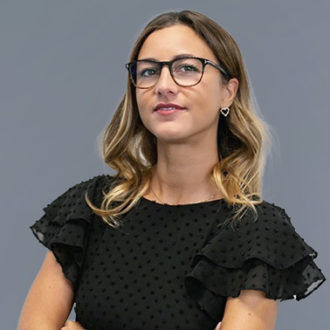 Donna bionda con occhiali in abito nero, braccia conserte, su sfondo grigio.