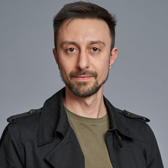 Uomo con giacca nera e maglietta verde oliva su sfondo grigio.