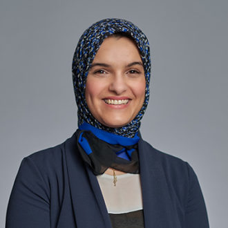 Donna sorridente con hijab blu e nero e blazer blu scuro su sfondo grigio.