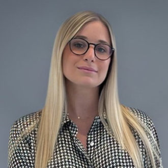 Donna bionda con occhiali e camicia a quadretti su sfondo grigio.