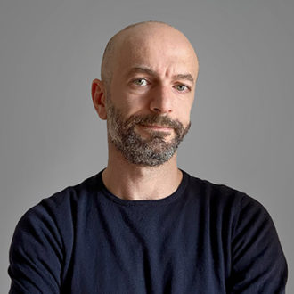 Uomo calvo con barba in maglia nera su sfondo grigio.