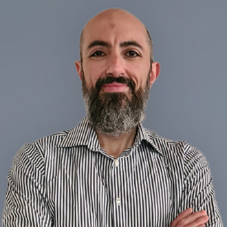 Persona con camicia a righe verticali e barba folta su sfondo grigio.