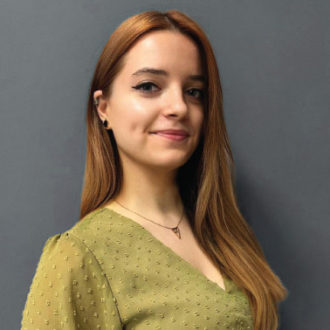 Giovane donna con capelli castano chiaro e vestito verde di fronte a sfondo grigio.